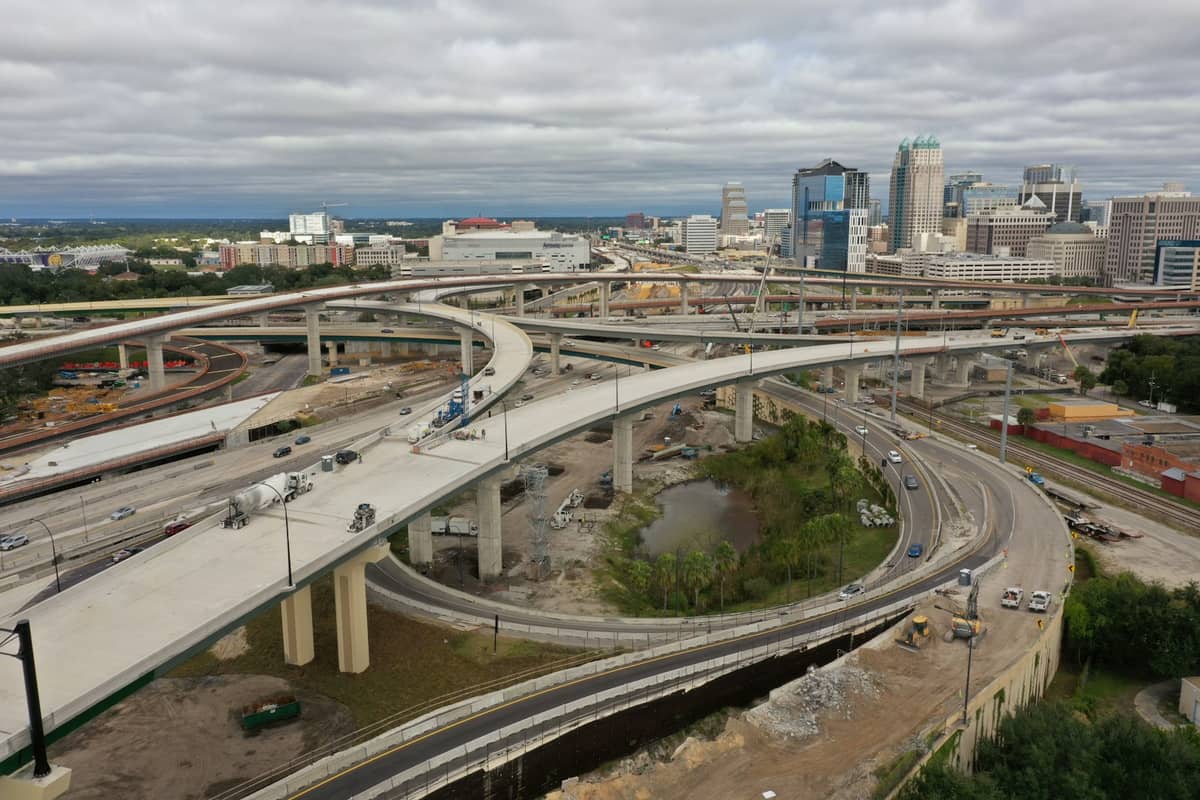 Aerial view of complex highway interchange with downtown Orlando skyline in the background under cloudy skies.