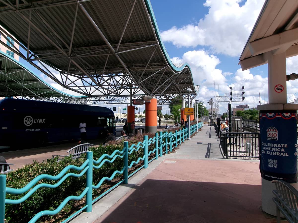 Covered transit center in Orlando with a LYNX bus at the platform, passengers waiting, and railroad tracks visible under a sunny sky.