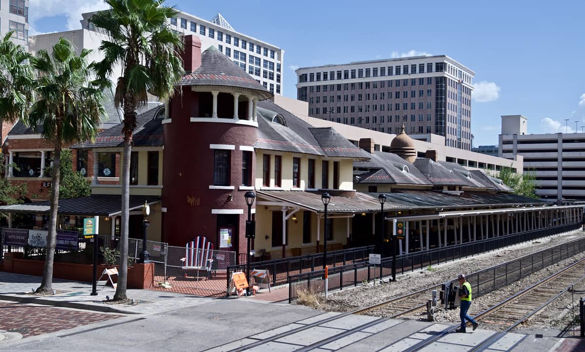 Historic downtown Orlando train station with palm trees, railroad tracks, and modern office buildings in the background.