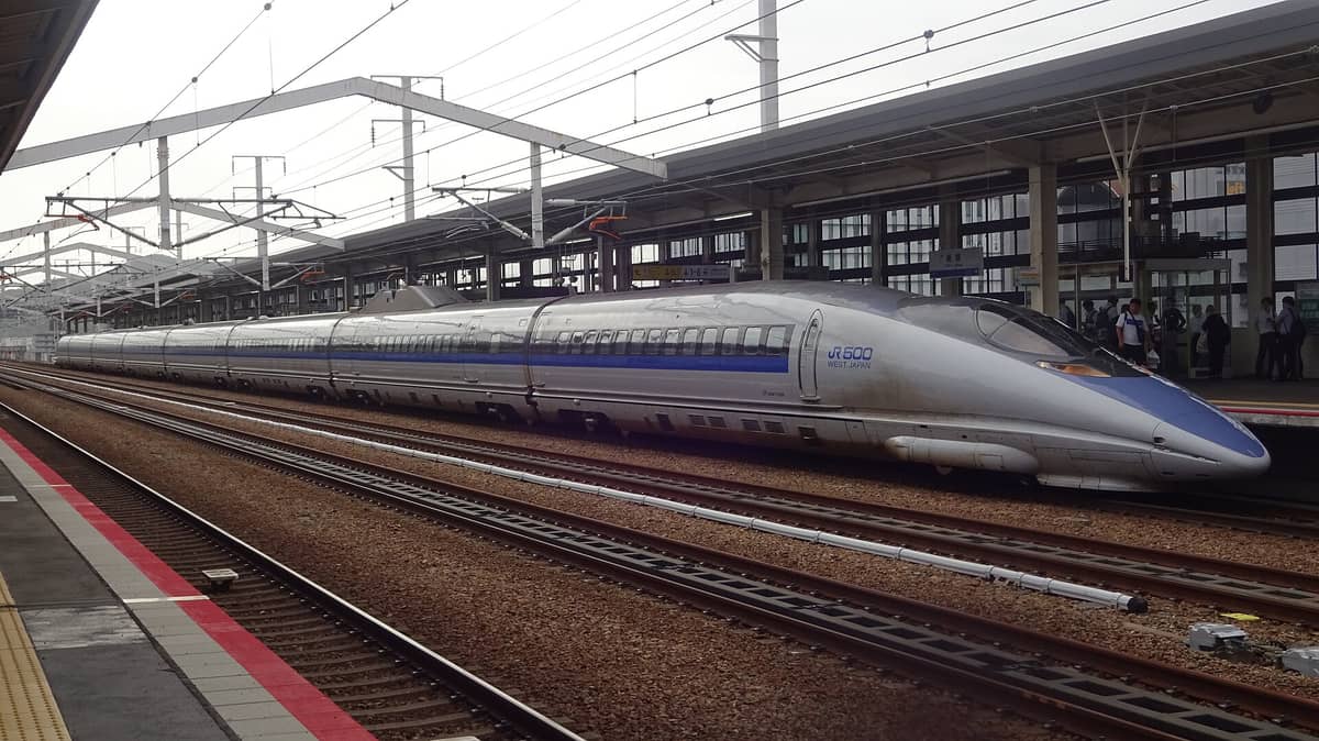A JR West 500 series shinkansen train at a platform, with several tracks and station structures in view.