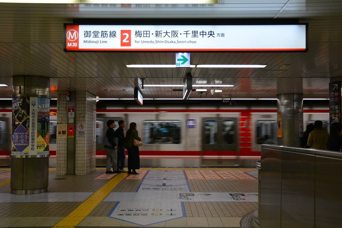 Osaka Metro Midosuji Line platform with red train, directional signs, and passengers waiting under bright lighting.