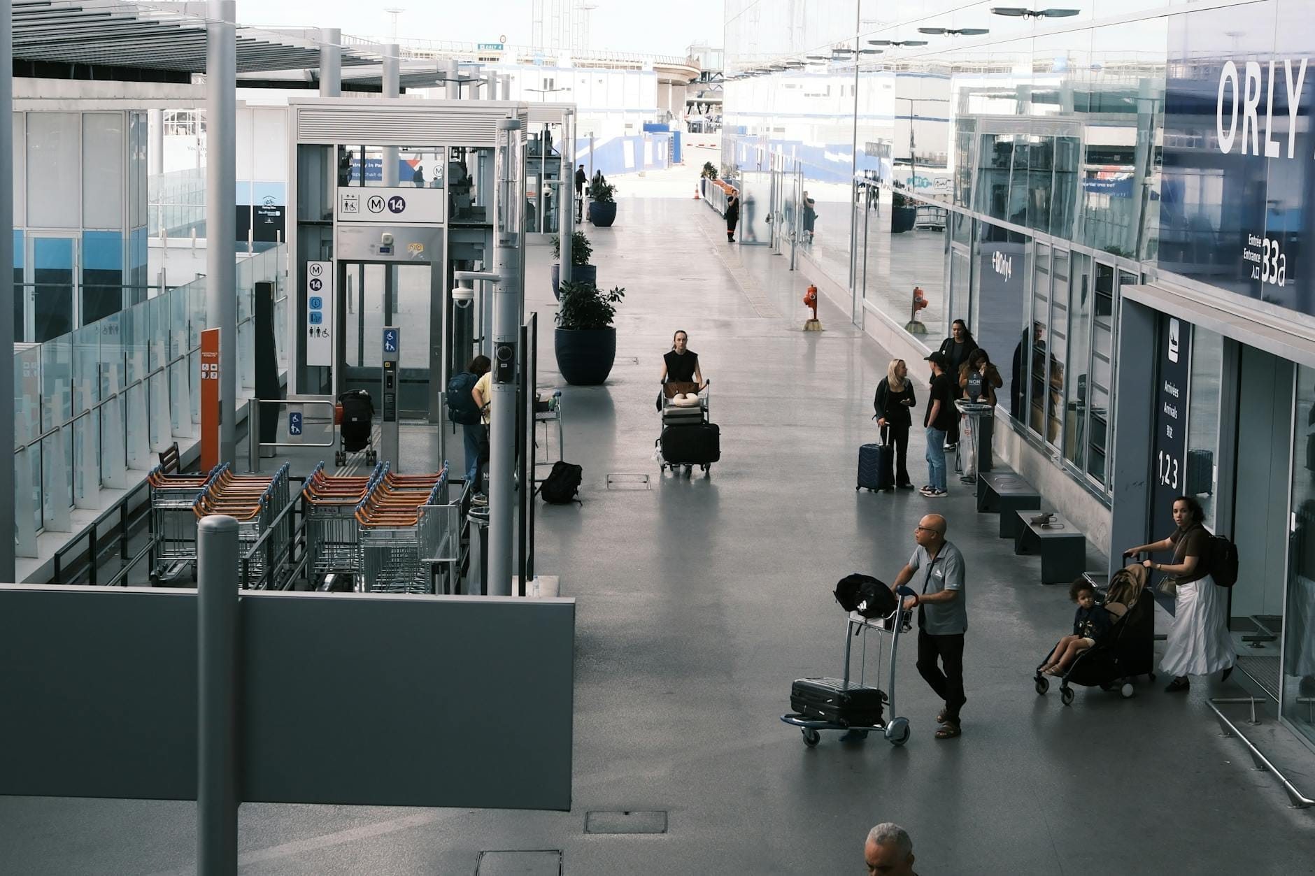 Außenansicht des Flughafens Paris Orly mit sichtbaren Personen, Gepäckwagen und gläsernem Terminaleingang, deutlich gekennzeichnet mit dem Schild 'ORY'.