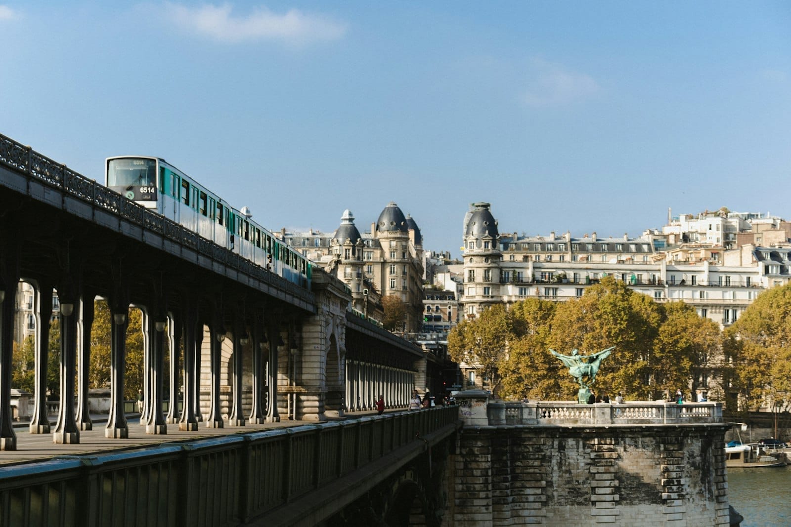 Un treno della Métro di Parigi attraversa il ponte di Bir-Hakeim sopra la Senna, con i classici edifici parigini e gli alberi sullo sfondo in una giornata limpida.
