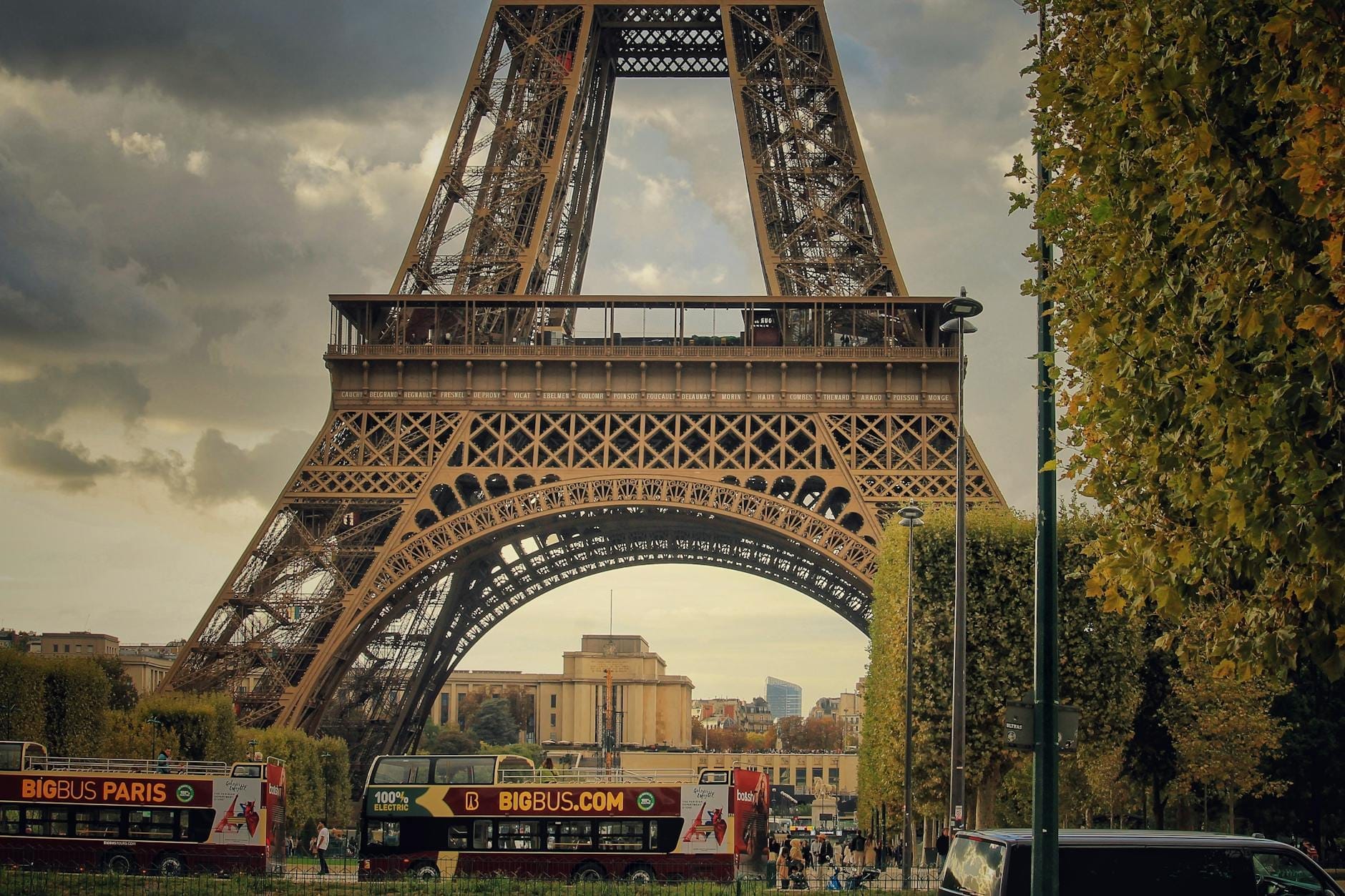 Zwei Stadtrundfahrtbusse in Paris fahren unter dem Eiffelturm auf einer Stadtstraße entlang, gesäumt von Bäumen.