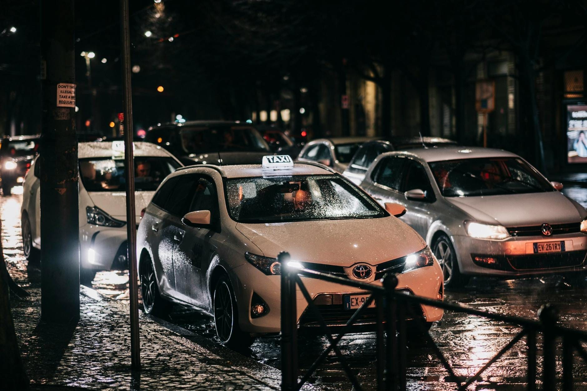 Weißes Taxi mit beleuchtetem Dachschild wartet im Verkehr auf einer regnerischen Pariser Straße bei Nacht, umgeben von anderen Autos.