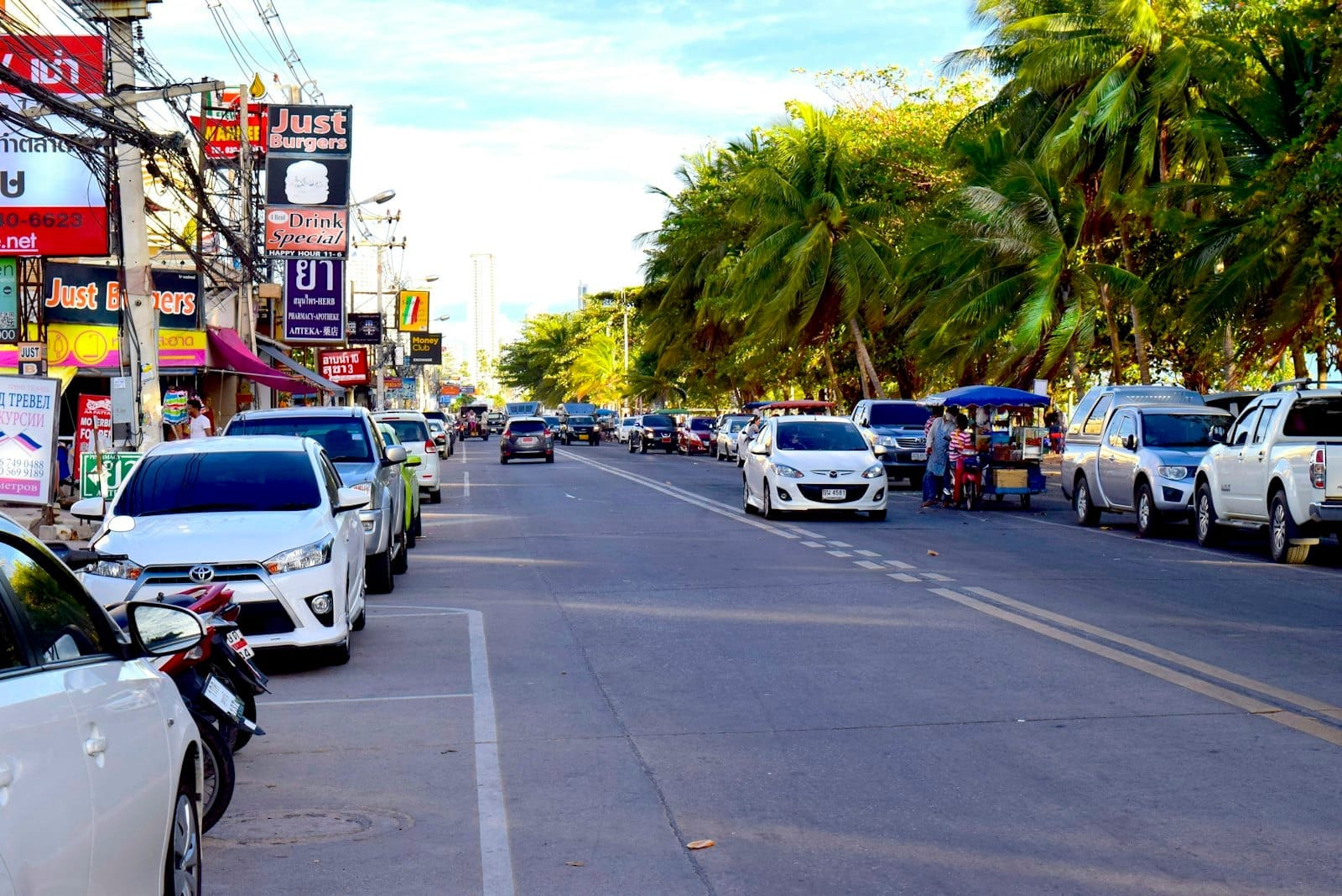 Rua movimentada de Pattaya com carros, veículos estacionados, palmeiras e lojas dos dois lados, capturando as opções de transporte e a atmosfera urbana da cidade.