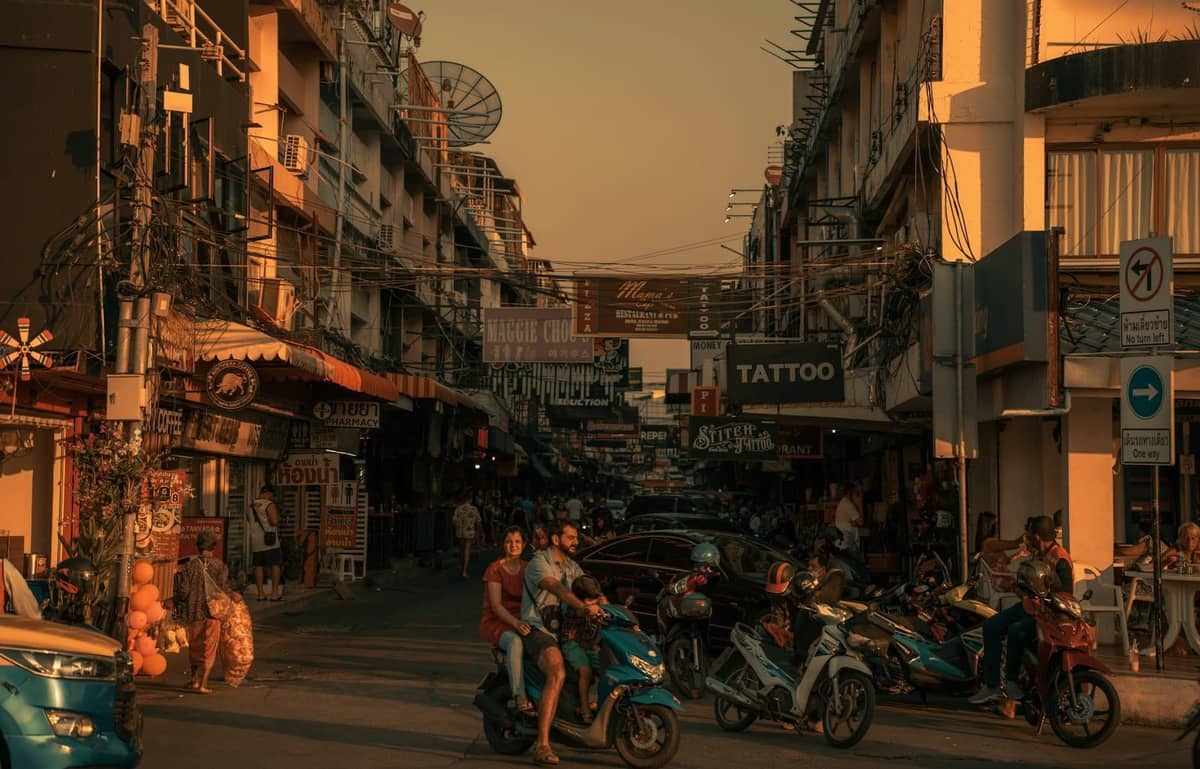 Busy Pattaya street at sunset with several people riding motorbikes and scooters, surrounded by shops and dense urban activity.