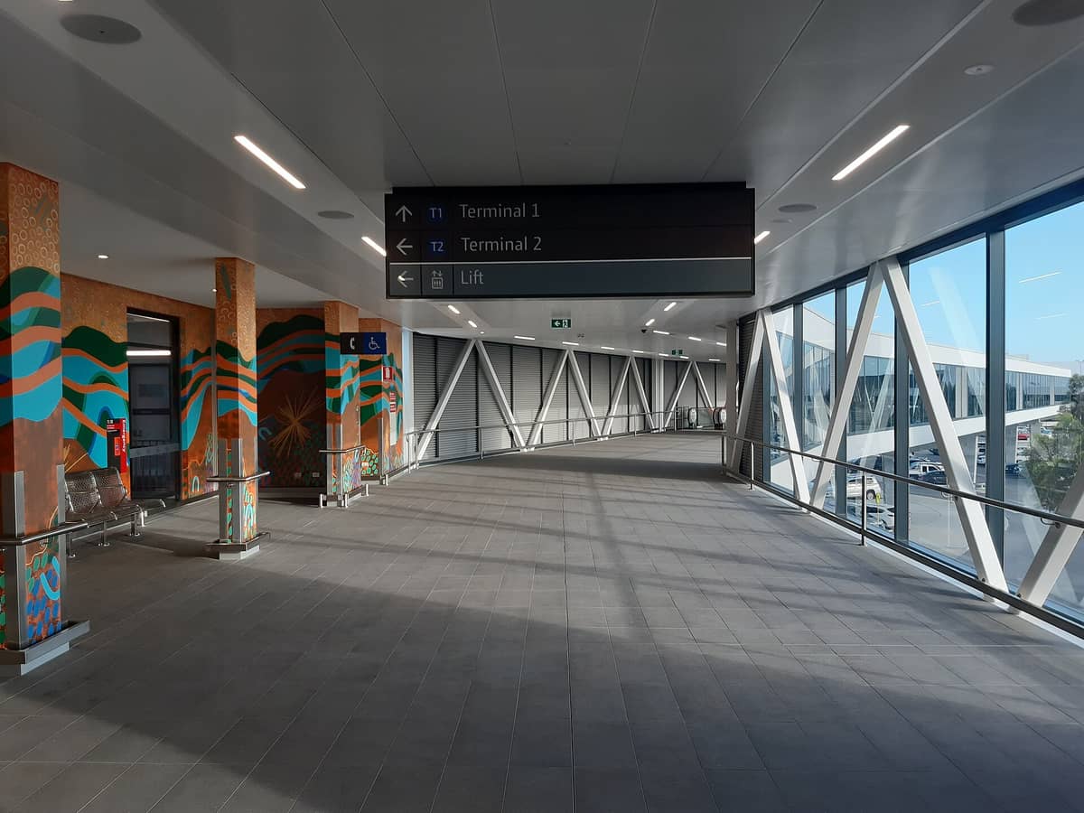 Moderne Skybridge am Flughafen Perth mit Hinweisschildern zu Terminal 1 und 2, großen Fenstern und bunten Wandgemälden an den Säulen.