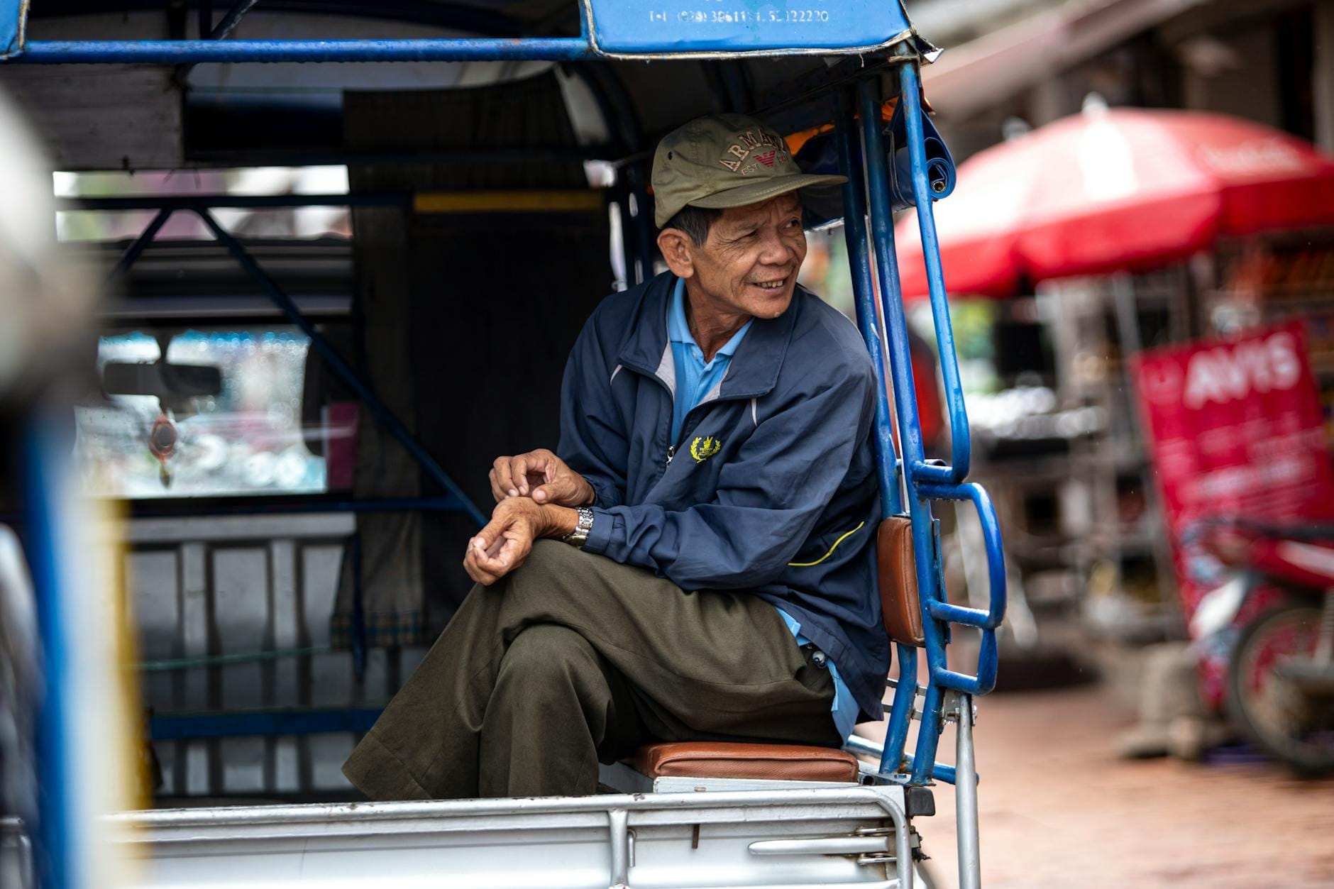 An older man sitting on the bench of a blue open-backed vehicle, likely a songthaew, in a street market setting in Thailand.