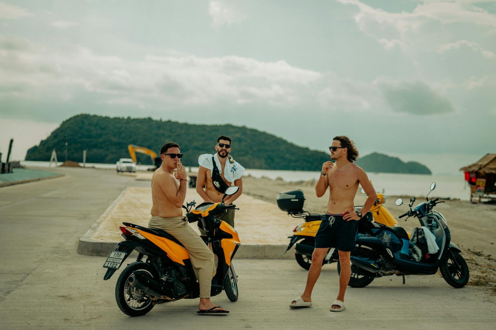 Three people with scooters near a beachside road with hills and ocean in the background, typical of Phuket's popular transport style.