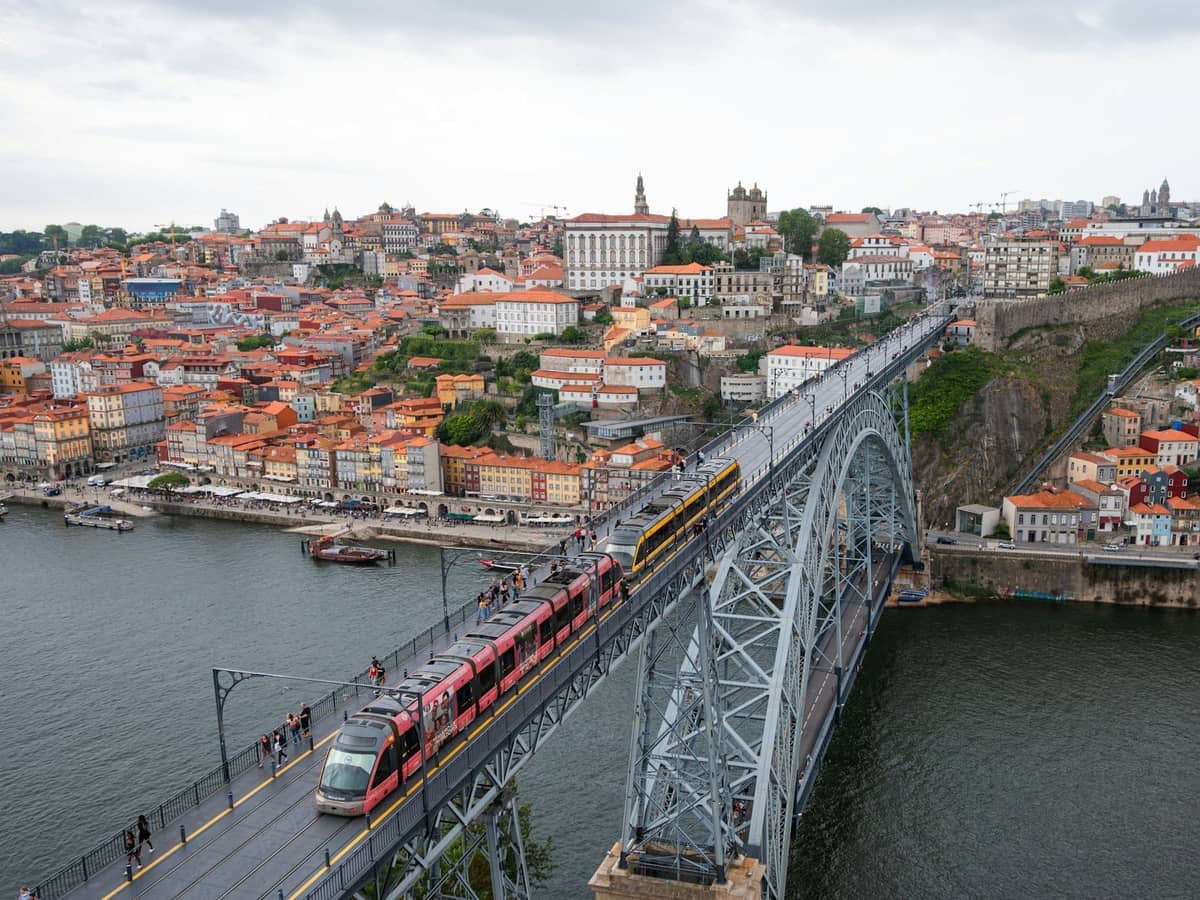 Trem modern dan trem kuning klasik melintasi Jembatan Dom Luís I di atas tepi sungai bersejarah Porto dengan cakrawala kota yang terlihat.