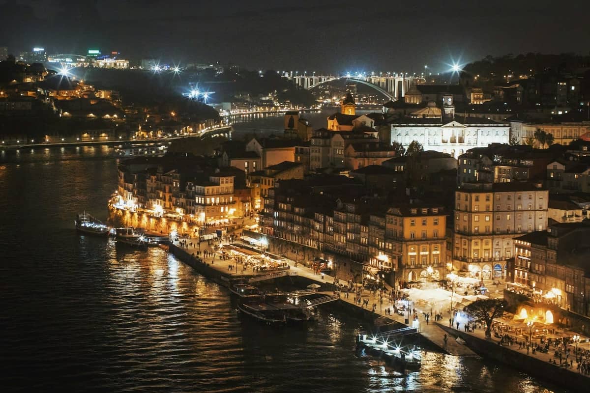 Pemandangan malam tepi sungai bersejarah Porto dengan gedung-gedung, lampu yang semarak, dan kota yang tercermin di sungai Douro.