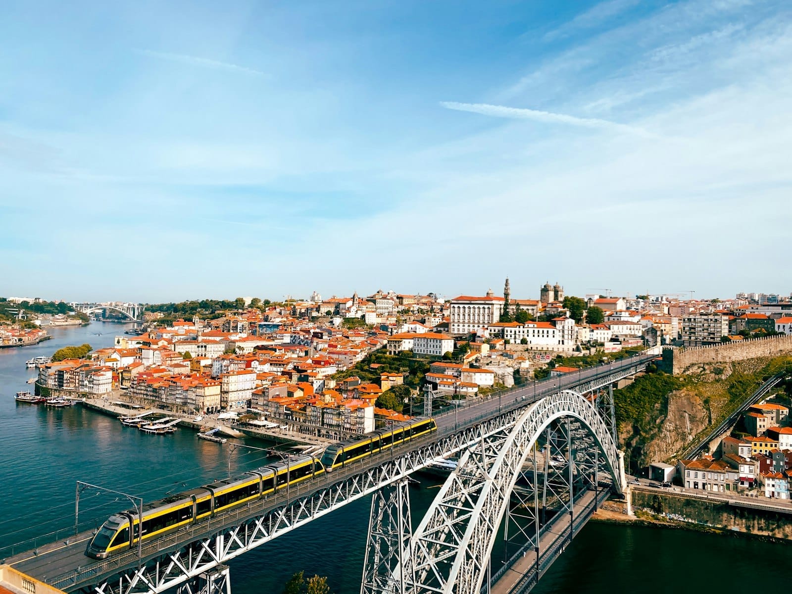 Vista panorâmica do Porto com a icônica Ponte Dom Luís I, um trem amarelo do metrô cruzando sobre o Rio Douro e o histórico skyline da cidade ao sol.