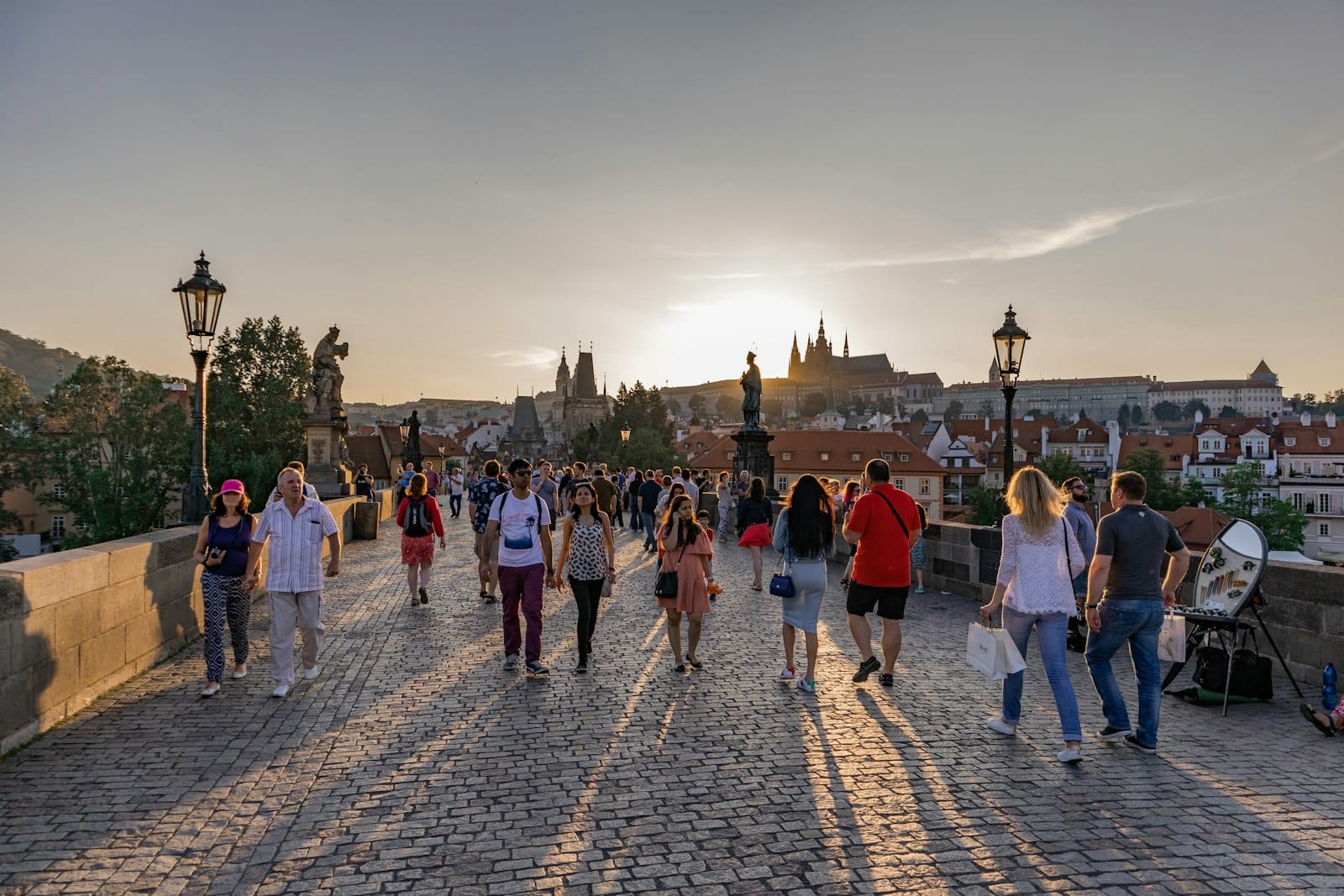 Menschen, die bei Sonnenuntergang über die historische Karlsbrücke in Prag spazieren, mit dem Prager Schloss in der Ferne.