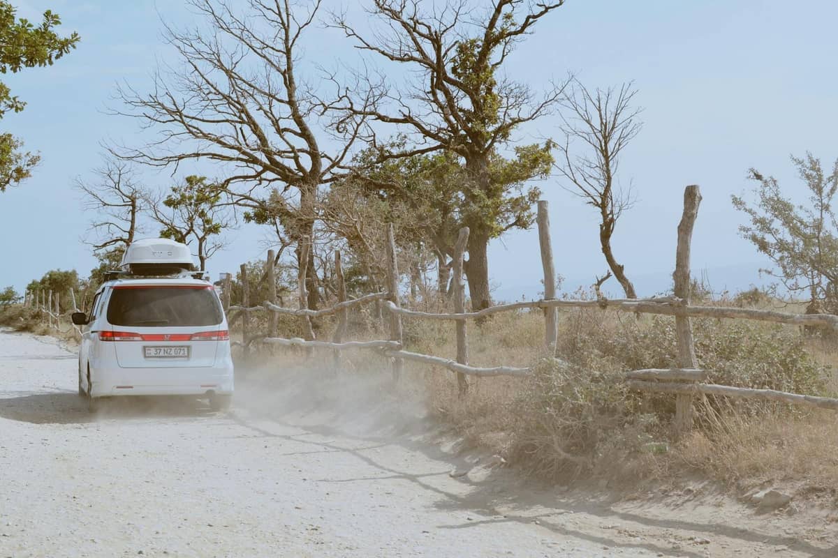 Car driving on a dusty rural road lined with rustic wooden fences and sparse trees under a bright sky.