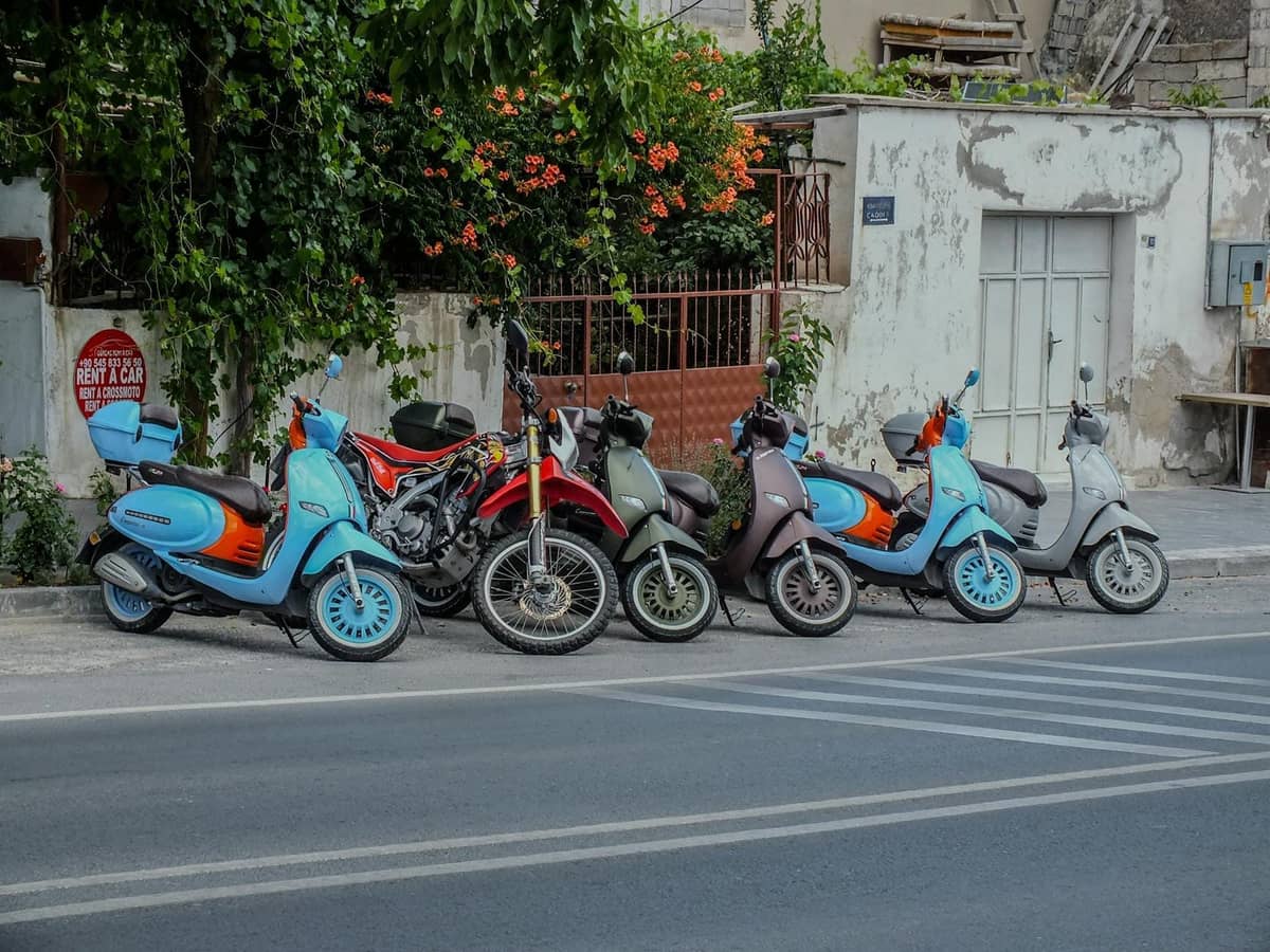 Several colorful scooters and a motorcycle parked by the roadside in a Greek village with whitewashed buildings and flowers.