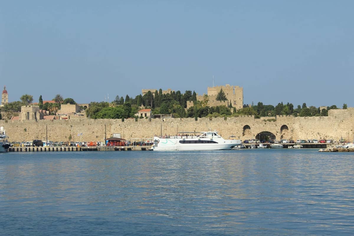 A white passenger ferry arrives at Rhodes Port with the medieval city walls and towers in the background on a clear day.