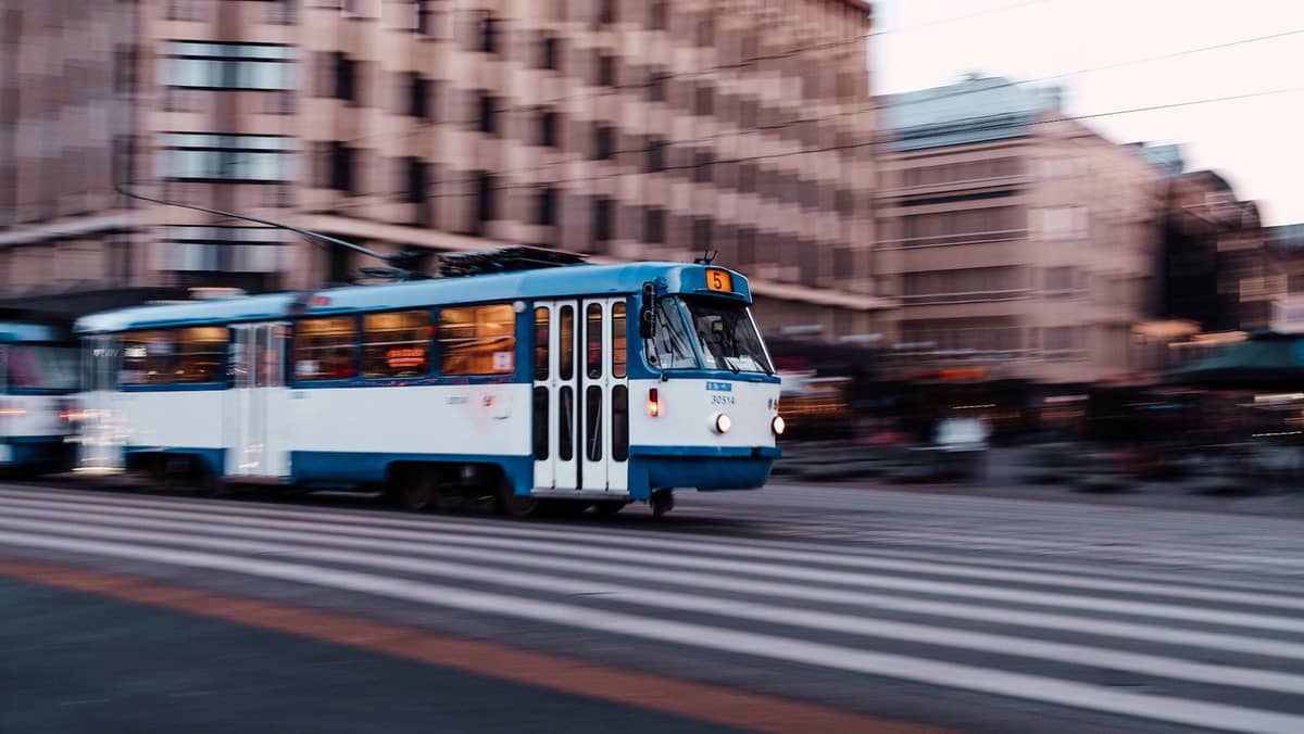 Eine blau-weiße Straßenbahn fährt durch eine Straße im Zentrum von Riga, im Hintergrund sind Stadtgebäude und Geschäfte zu sehen.