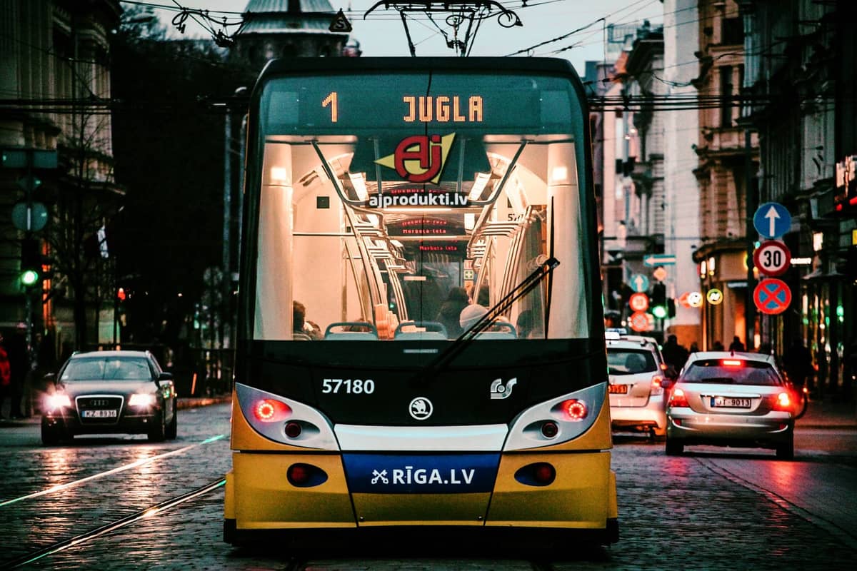 Frontansicht einer Rigaer Straßenbahn mit den Aufschriften 'Jugla' und 'Riga.lv' auf einer Stadtstraße in der Abenddämmerung mit Autos und beleuchteten Gebäuden ringsum.