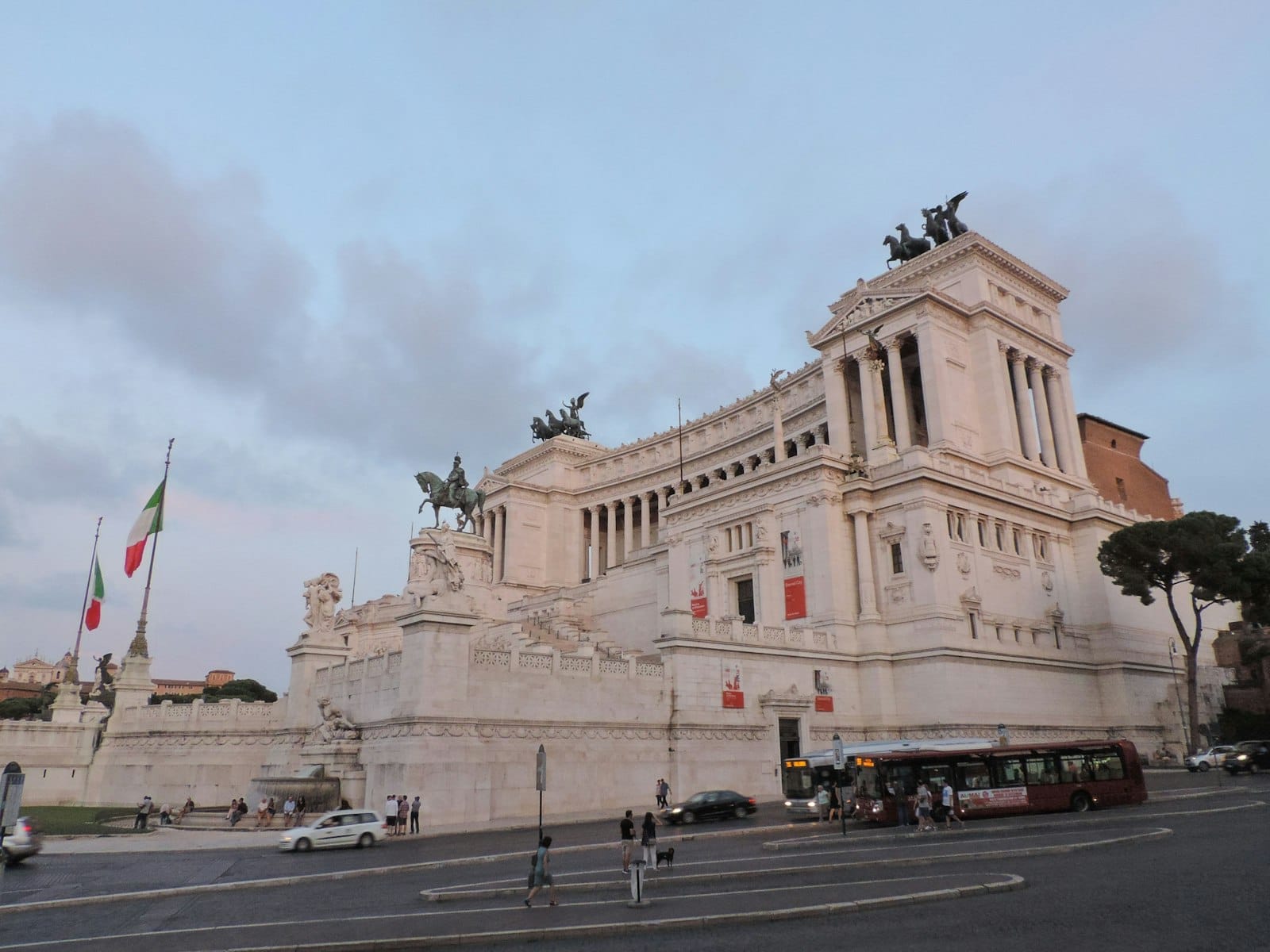 A Rome city bus at a stop in front of the Vittoriano monument, with cars and people nearby in early evening light.