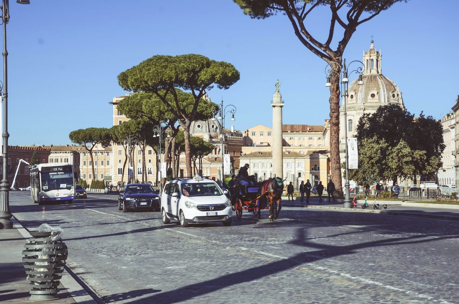 Wide city street in Rome with cars, taxis, a bus, and a horse carriage, with domed churches and Roman pine trees in the background under a clear sky.