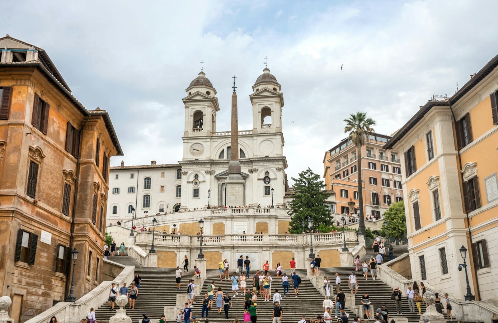 Wide view of the Spanish Steps in Rome with people walking, surrounded by historic buildings under a cloudy sky.