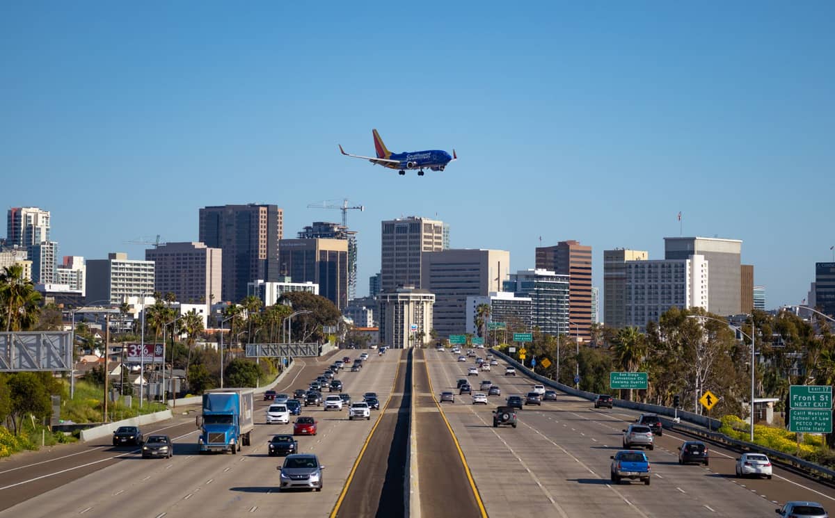 Eine breite Stadtautobahn in San Diego führt auf die Innenstadt mit ihren Wolkenkratzern zu, ein Flugzeug im Landeanflug und verschiedene Fahrzeuge auf der Straße unter einem strahlend blauen Himmel.