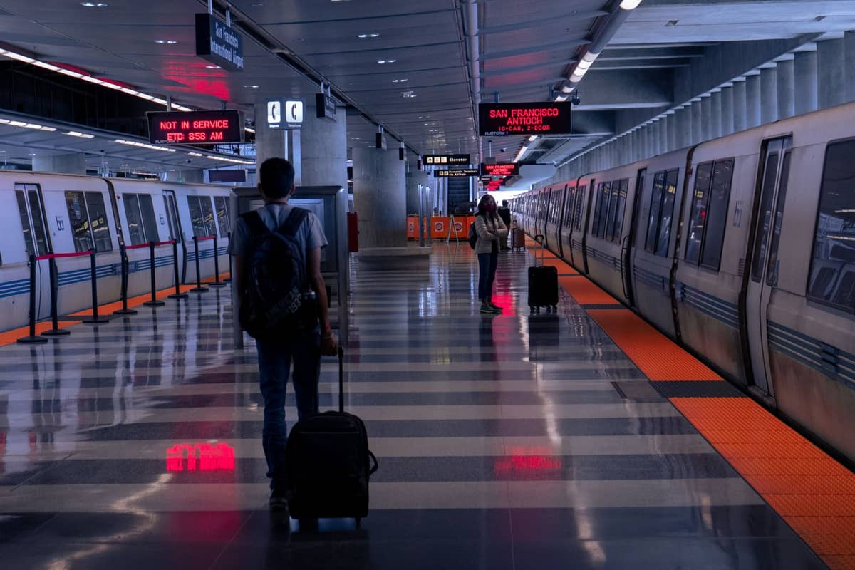 Menschen mit Gepäck, die auf einem sauberen, modernen BART-Bahnsteig mit zwei BART-Zügen und elektronischen Anzeigen darüber spazieren.