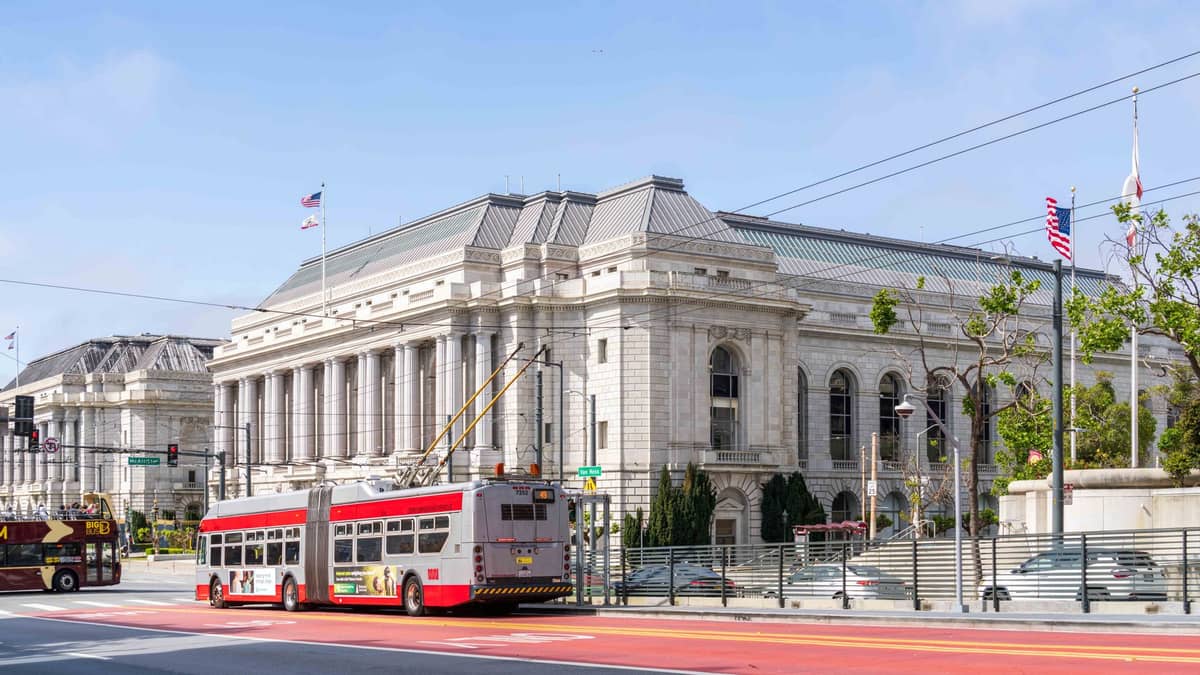 Ein rot-weißer Muni-Oberleitungsbus vor dem War Memorial Opera House in San Francisco, mit sichtbaren Oberleitungsdrähten und klaren Stadtverkehrsmarkierungen.