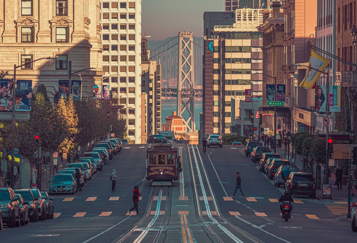 San Francisco Cable Car, der bei Sonnenuntergang eine steile Straße hinaufährt, mit Stadtgebäuden und der Bay Bridge im Hintergrund.