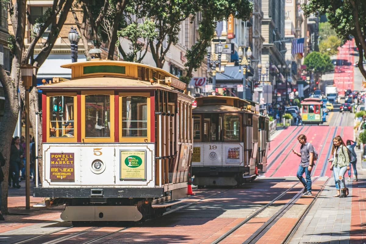 Dos teleféricos clásicos de San Francisco suben y bajan por una concurrida calle de la ciudad mientras los peatones cruzan, mostrando las opciones de transporte público local.