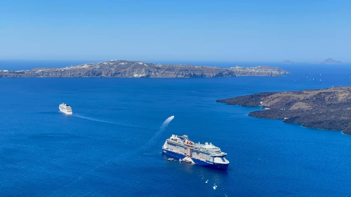 Luftaufnahme der Caldera von Santorini mit mehreren Fähren und Kreuzfahrtschiffen, die über blaues Wasser und Vulkaninseln anlegen.