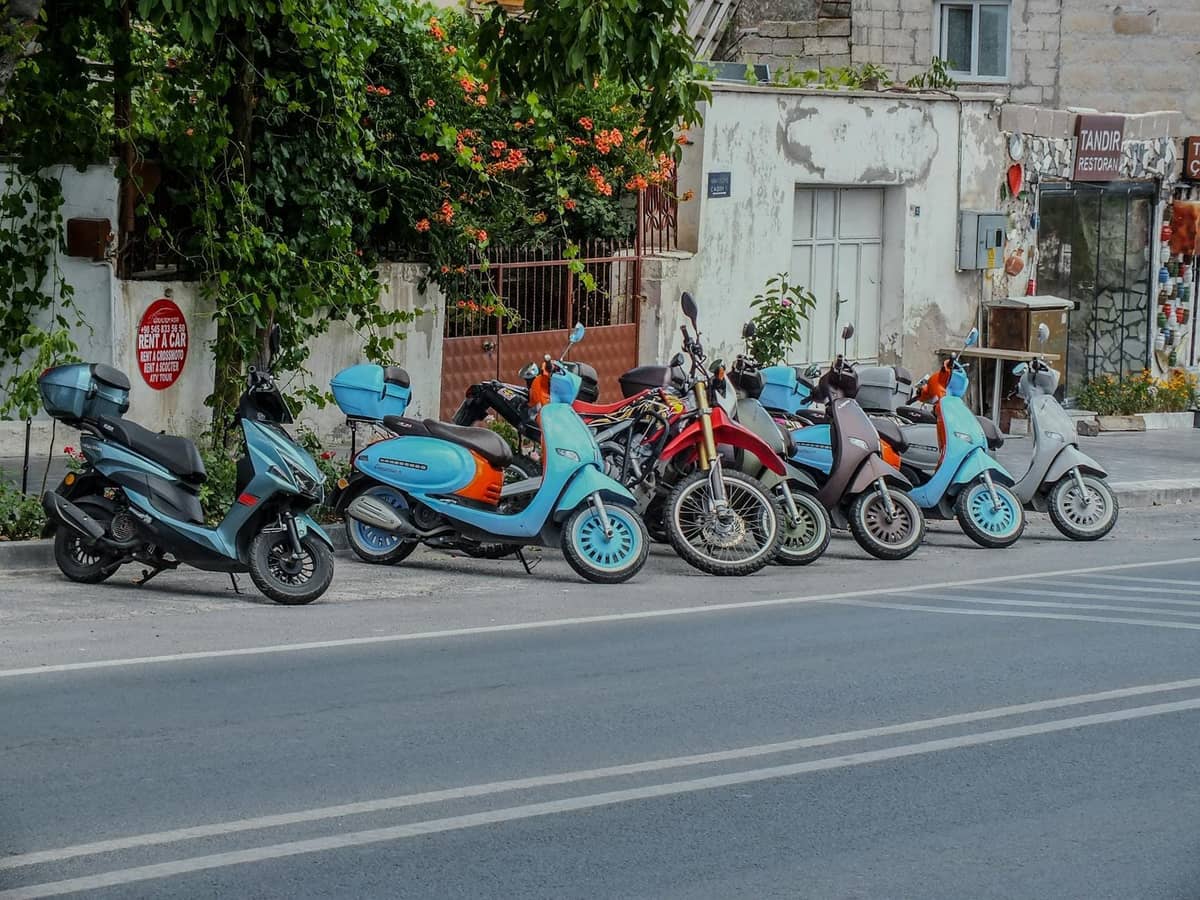 Reihe von Scootern und einem Motorrad, die in einer Straße eines mediterranen Dorfes mit blühenden Pflanzen und weiß getünchten Gebäuden geparkt sind.