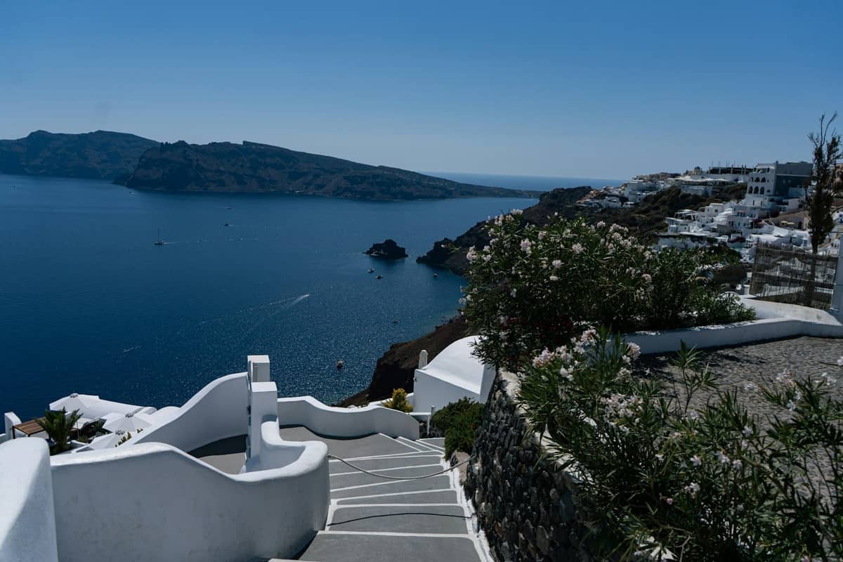Ein malerischer Fußweg mit weißen Treppen und Geländern, der sich entlang der Santorini-Caldera schlängelt und Ausblicke auf das blaue Meer, Klippen und entfernte Dörfer bietet.