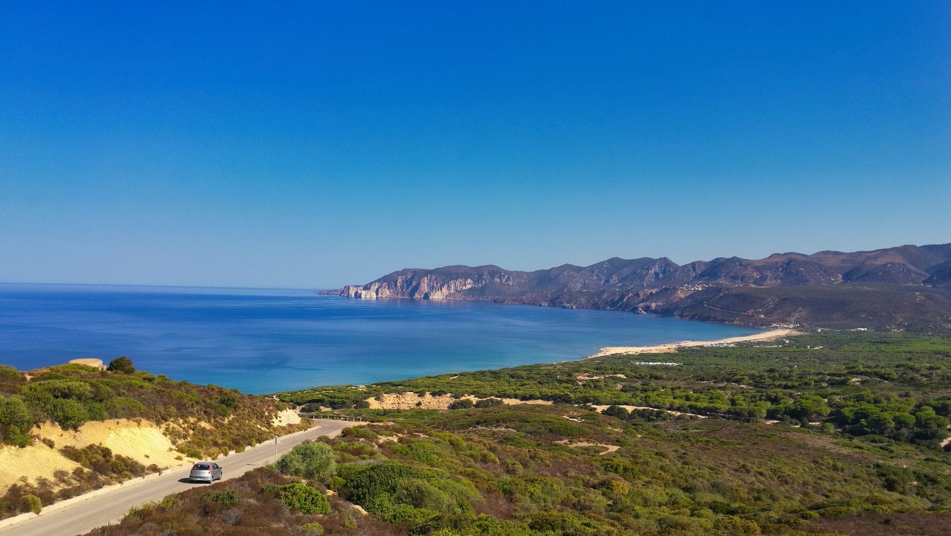 Auto fährt auf einer Küstenstraße auf Sardinien mit malerischen Klippen, grünen Hügeln und blauem Meer im Hintergrund unter einem klaren Himmel.