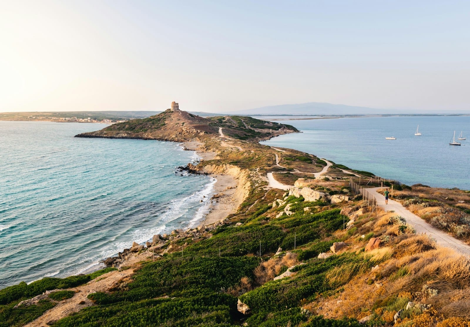 Panoramablick auf eine Küstenstraße, die sich entlang der zerklüfteten Küste Sardiniens windet – mit türkisfarbenem Wasser, Sandstränden und einem Turm auf einem Vorgebirge unter klarem Himmel.
