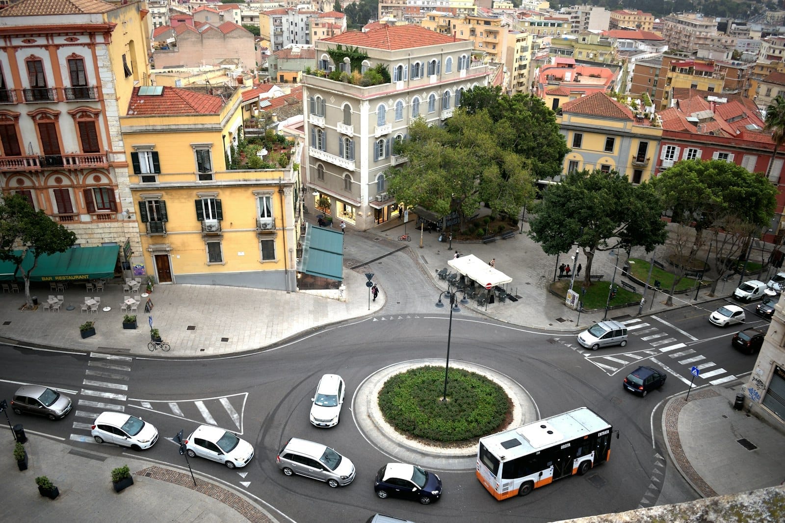 Ein Kreisverkehr in einem sardischen Stadtzentrum mit Taxis, Autos und einem Bus, die durch die Straßen fahren, umgeben von bunten historischen Gebäuden.
