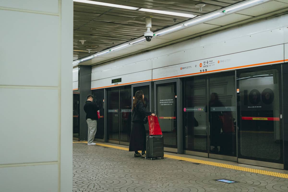 Pessoas esperando em uma plataforma dentro de uma estação de metrô de Seul com portas de plataforma de vidro e sinalização em coreano acima.