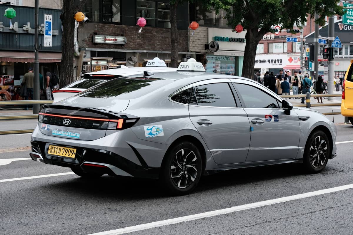 Um táxi Hyundai prateado com placas de táxi de Seul e placa de licença coreana circulando por uma rua movimentada em Seul com placas de lojas em coreano e vida urbana visível.