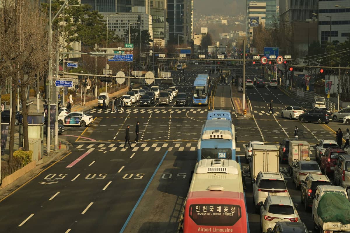 Vista ampla de uma grande avenida de Seul com ônibus municipais azuis, um ônibus Airport Limousine, táxis e trânsito intenso, emoldurada por arranha-céus modernos e letreiros em coreano.
