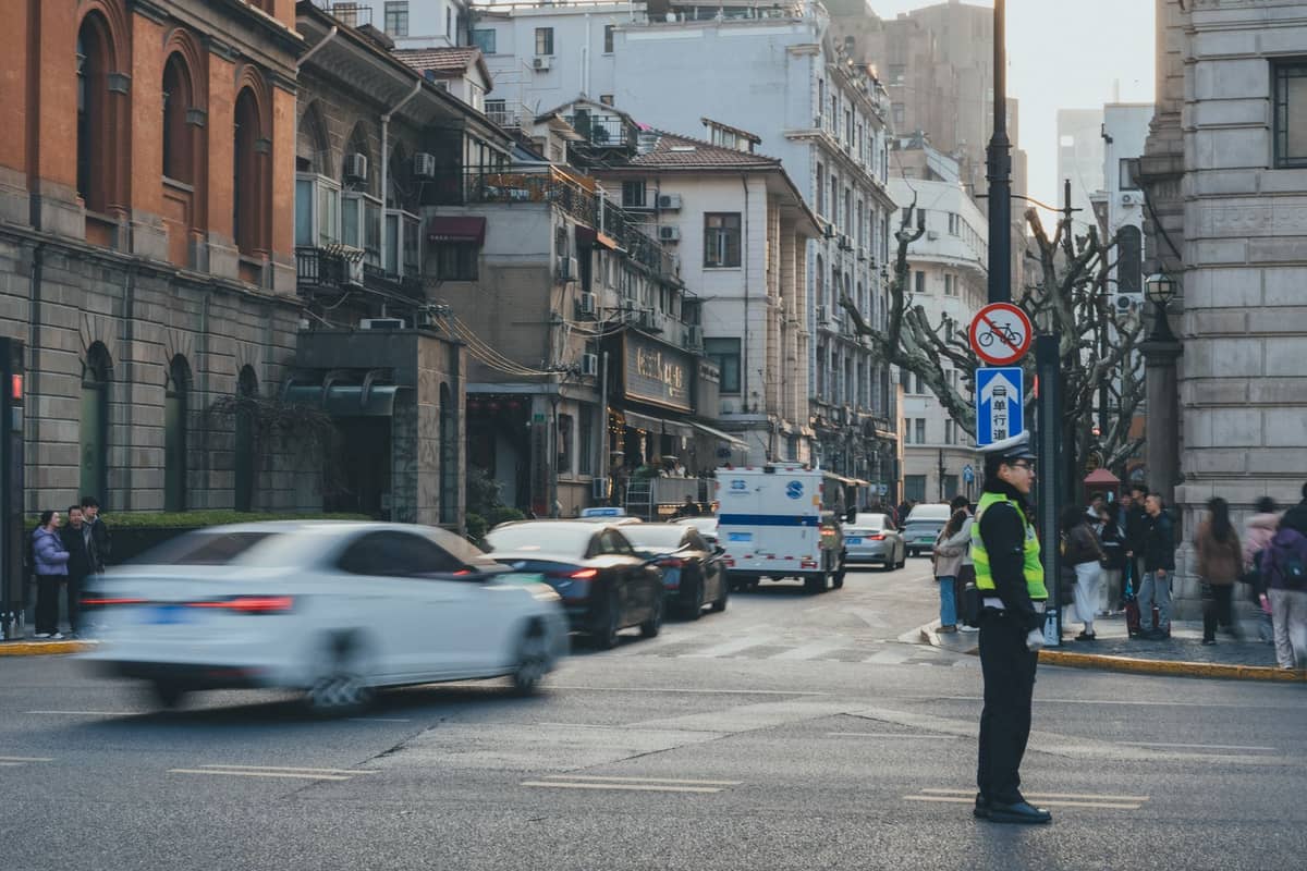 Eine Straßenszene in Shanghai mit mehreren Autos, einem Polizisten, der den Verkehr regelt, und historischen Stadtgebäuden im Hintergrund.