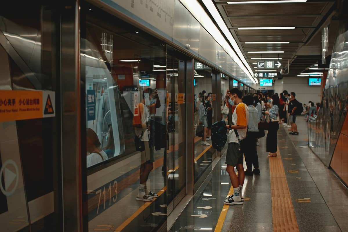 Passageiros aguardando ao lado de um trem moderno do Metrô de Shenzhen atrás de portas de vidro em uma plataforma de metrô limpa e bem iluminada.