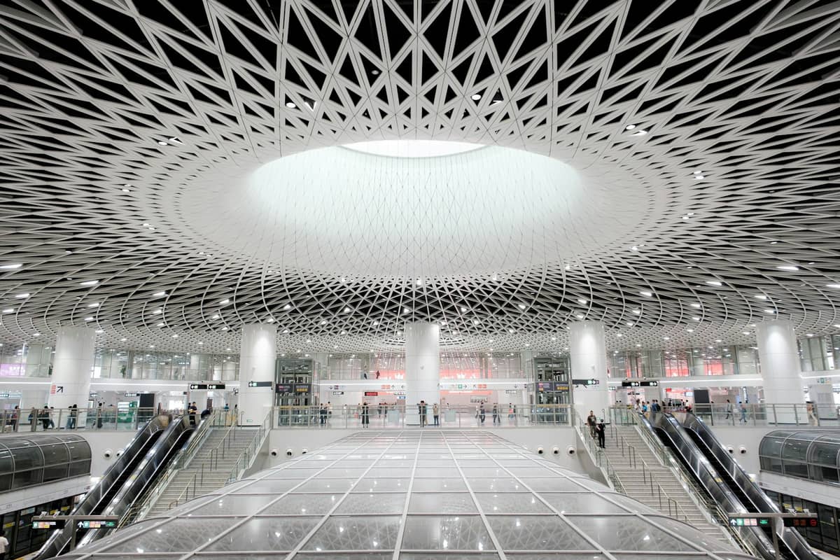 Vista ampla do interior moderno de uma estação de metrô de Shenzhen com o teto em cúpula treliçada, escadas rolantes e pessoas circulando pelo terminal.