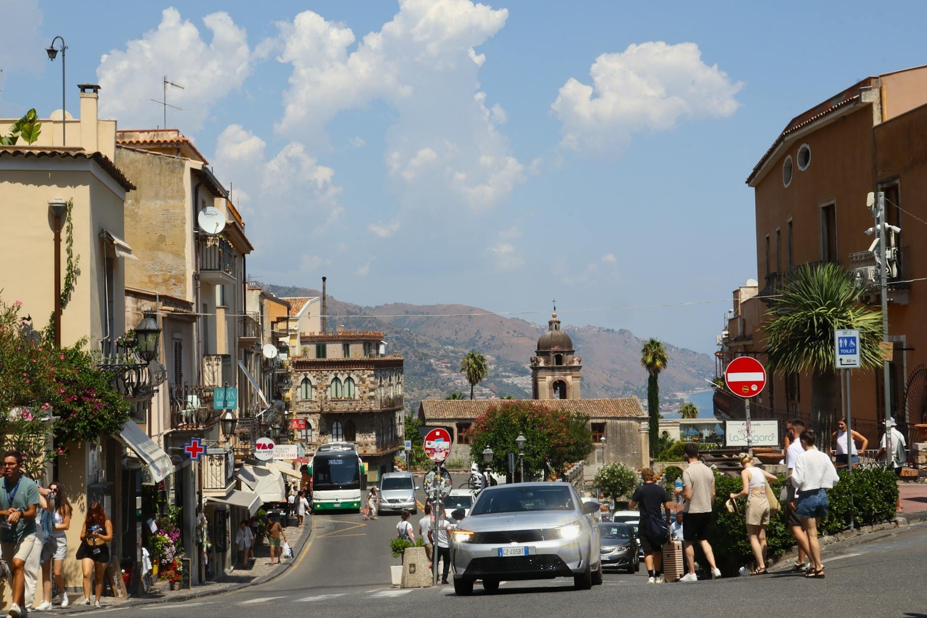 Eine lebhafte sizilianische Straße mit Autos, Bussen und Fußgängern, bunten historischen Gebäuden, Bergen im Hintergrund und mediterranem Flair.
