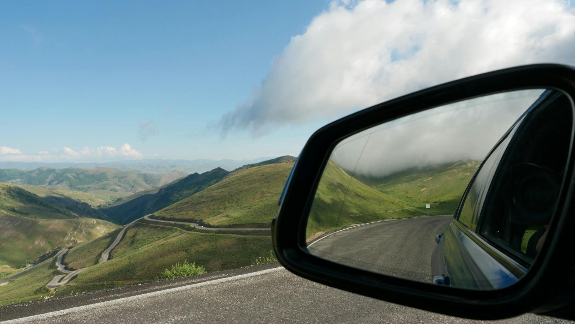 Blick aus dem Seitenspiegel eines Autos auf kurvenreiche Bergstraßen und grüne Hügel unter einem teils bewölkten Himmel in Sizilien.
