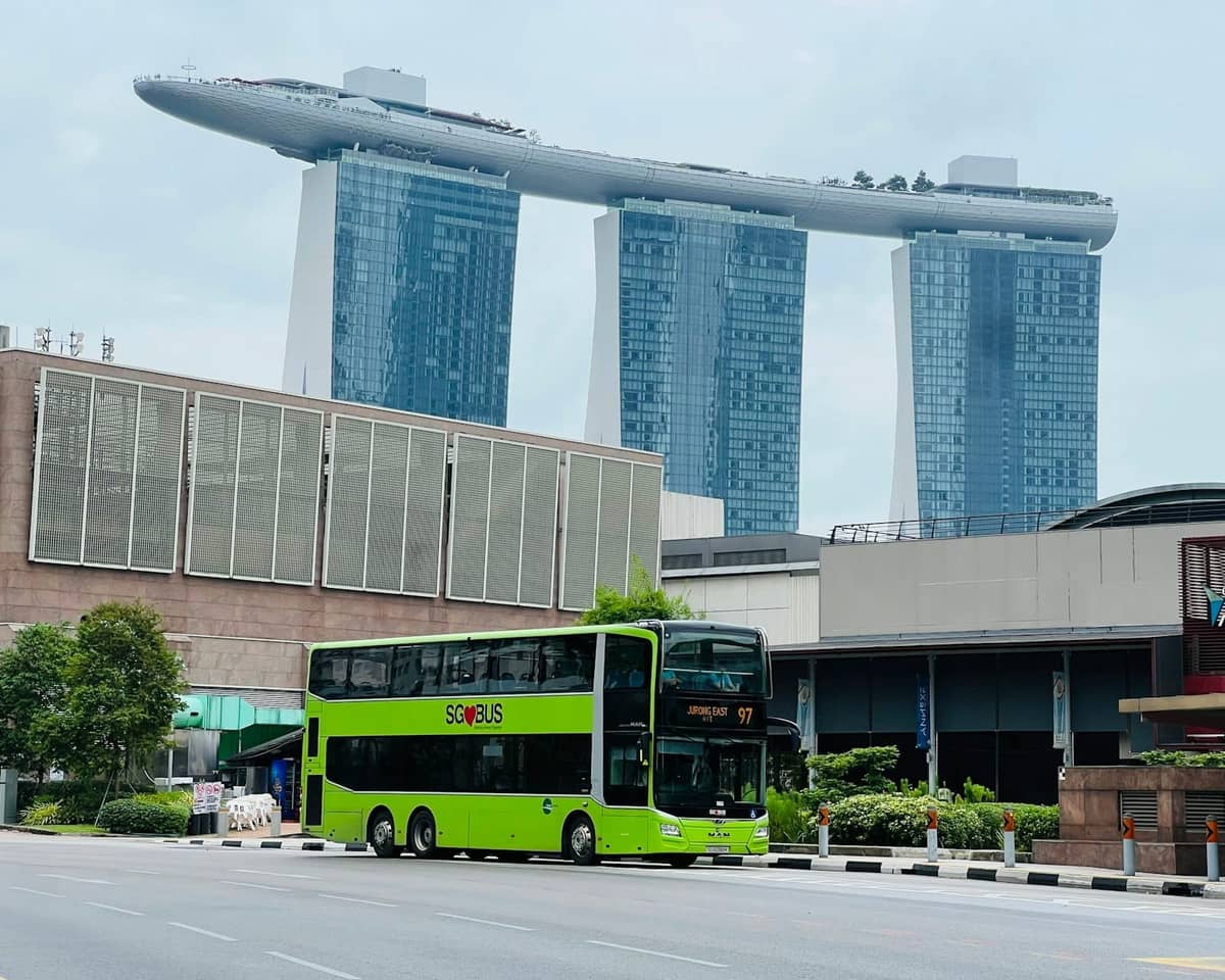 A bright green double-decker Singapore bus in front of the Marina Bay Sands hotel, clear sky and city buildings in background.