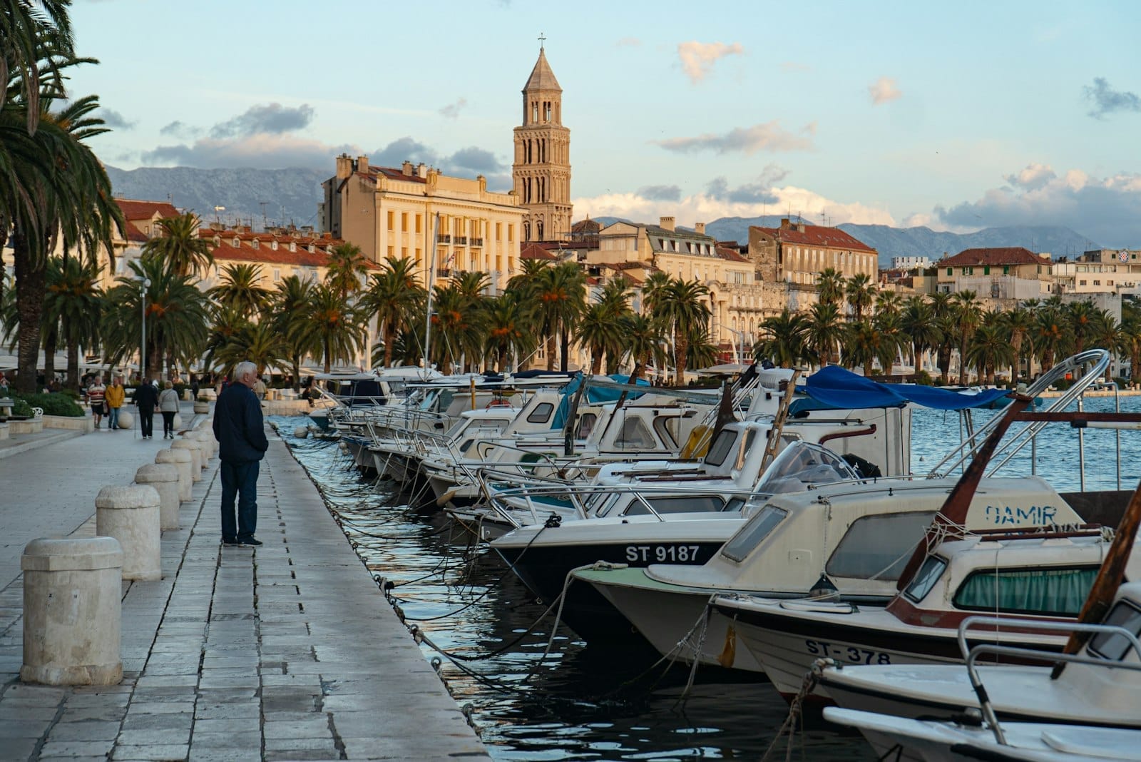 Barcos atracados ao longo do passeio Riva de Split, com palmeiras e o campanário do Palácio de Diocleciano ao fundo ao pôr do sol.