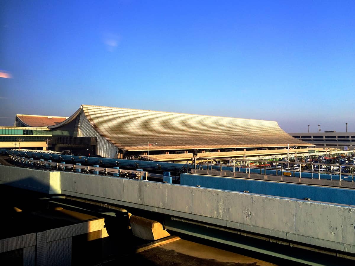 Außenansicht des Terminalgebäudes des Taiwan Taoyuan International Airport mit Parkplätzen und Transportspuren davor unter einem klaren blauen Himmel.