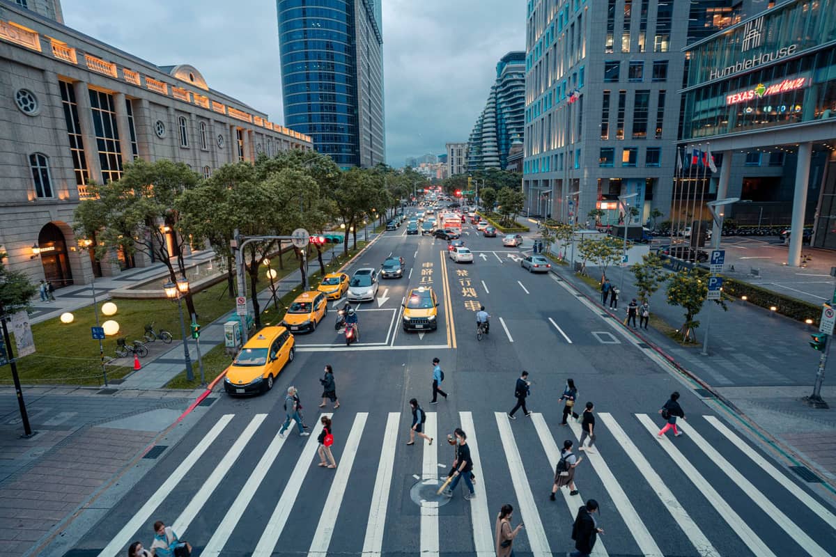 Mehrere gelbe Taxis warten an einem belebten Fußgängerüberweg in Taipei, während Fußgänger die Straße überqueren, umgeben von modernen Bürogebäuden und Stadtlichtern in der Abenddämmerung.