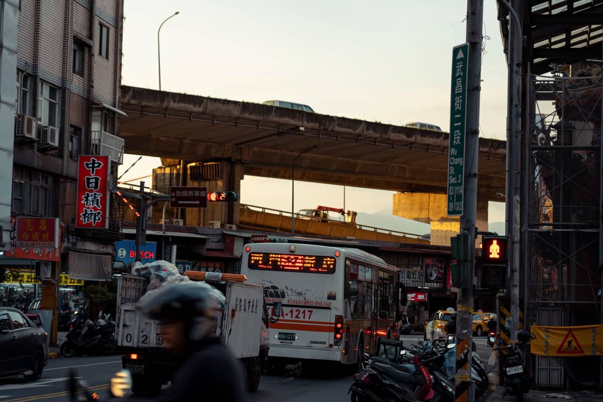 Stadtstraßenszene in Taipei bei Sonnenuntergang mit einem Stadtbus, Rollern und Gebäuden unter einer Überführung in einer städtischen Umgebung.